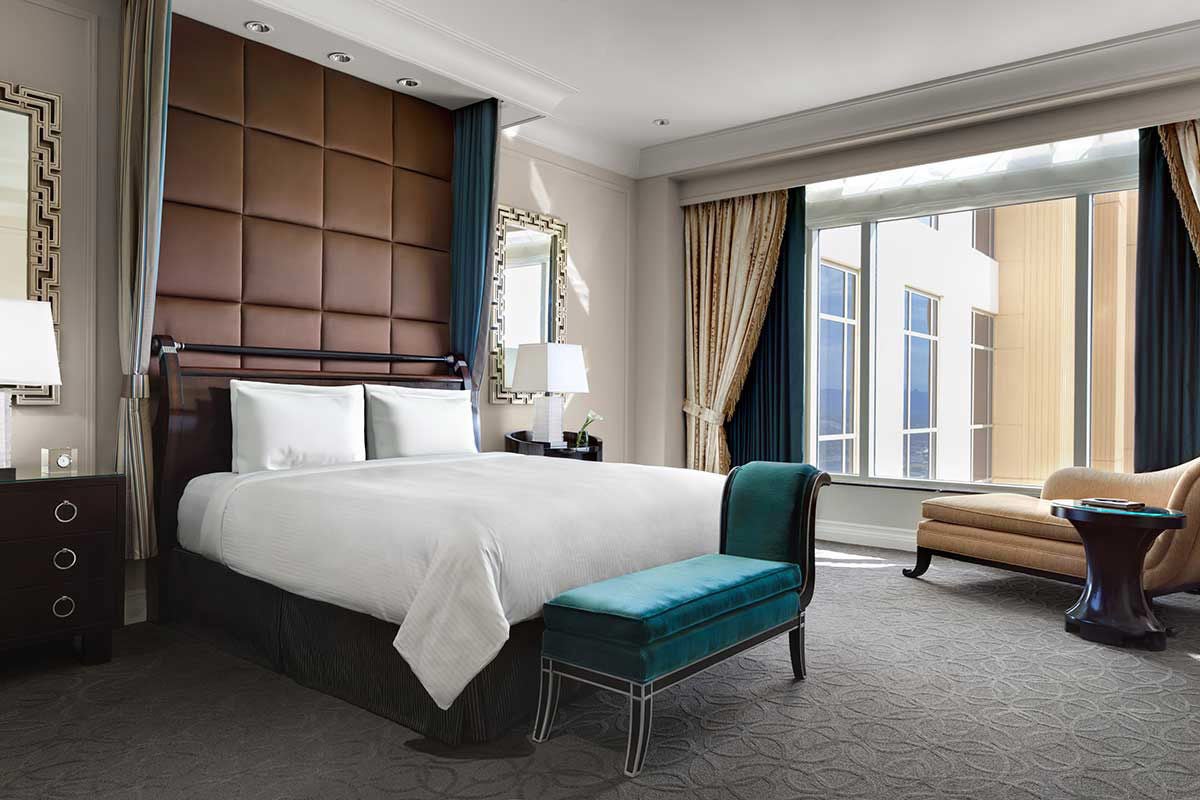 Hotel room with a King bed, chaise lounge in the right corner, bench at the end of the bed, two nightstands with a lamp each, and a large window on the right. 