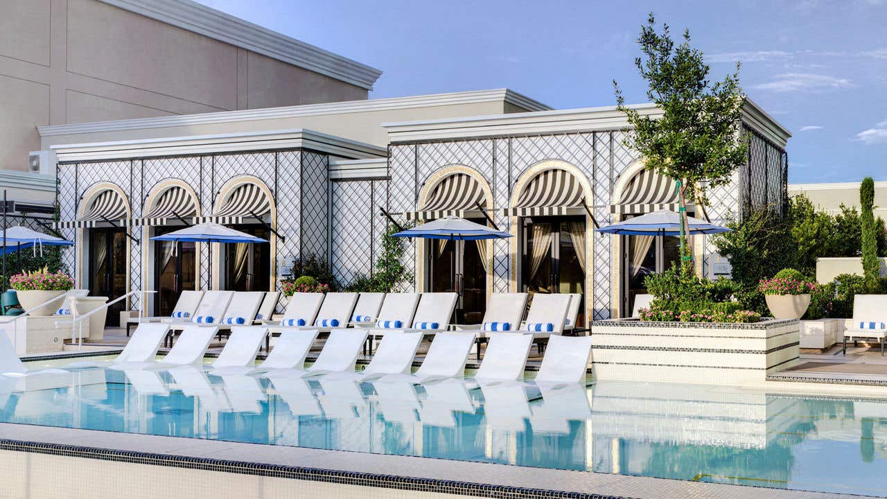 Row of white in water loungers in a shallow pool, positioned in front of cabanas with black and white striped awnings and surrounding greenery.