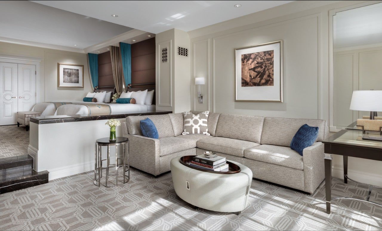 Elegant hotel room with double beds, modern decor, beige sofa, round ottoman, and a work desk under framed artwork.