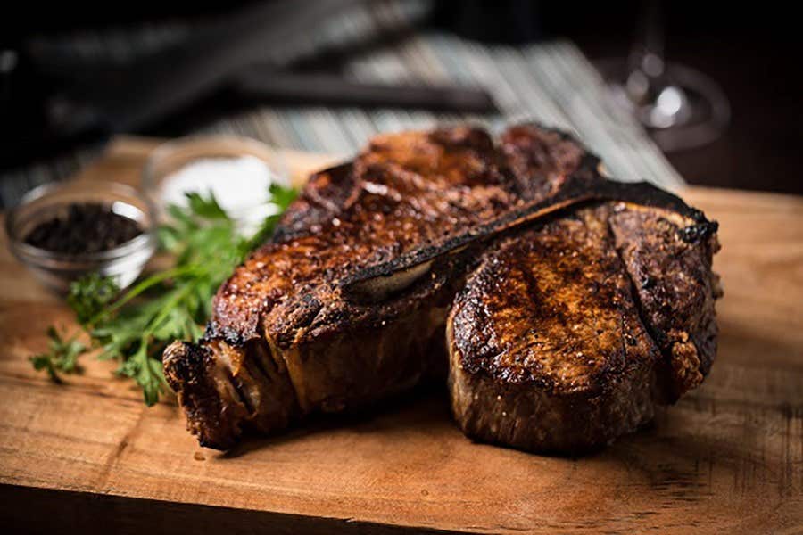 Juicy, grilled T-bone steak on a wooden board, garnished with herbs, and served with condiments in small bowls.