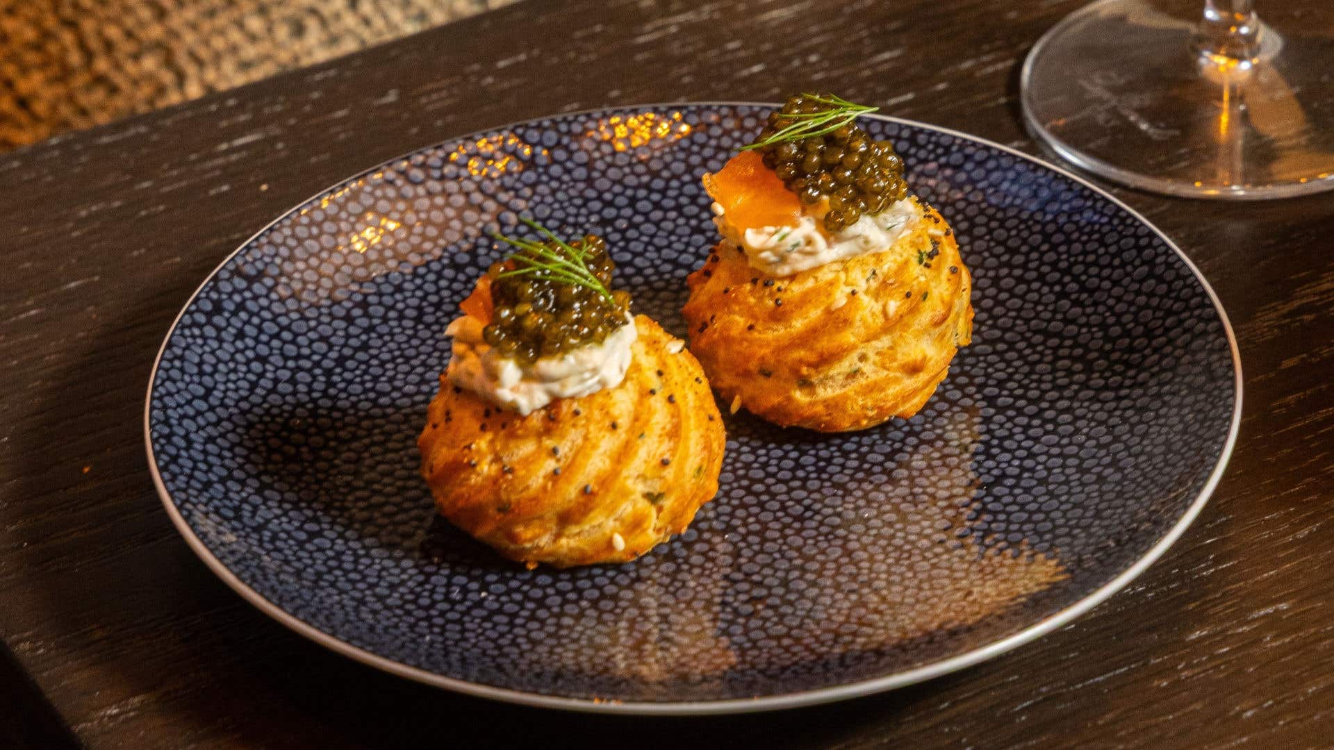 Two golden savory pastry puffs topped with cream, smoked salmon, caviar, and dill, served on a patterned blue plate.