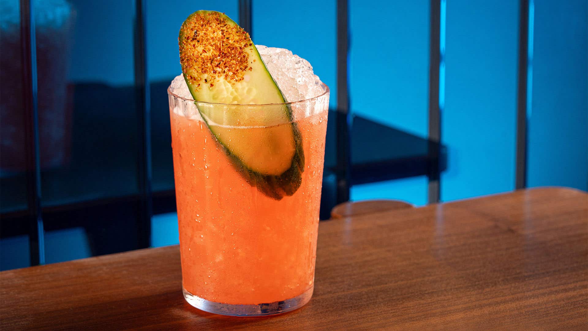Pink cocktail with ice and garnished with a cucumber slice with tajin. 