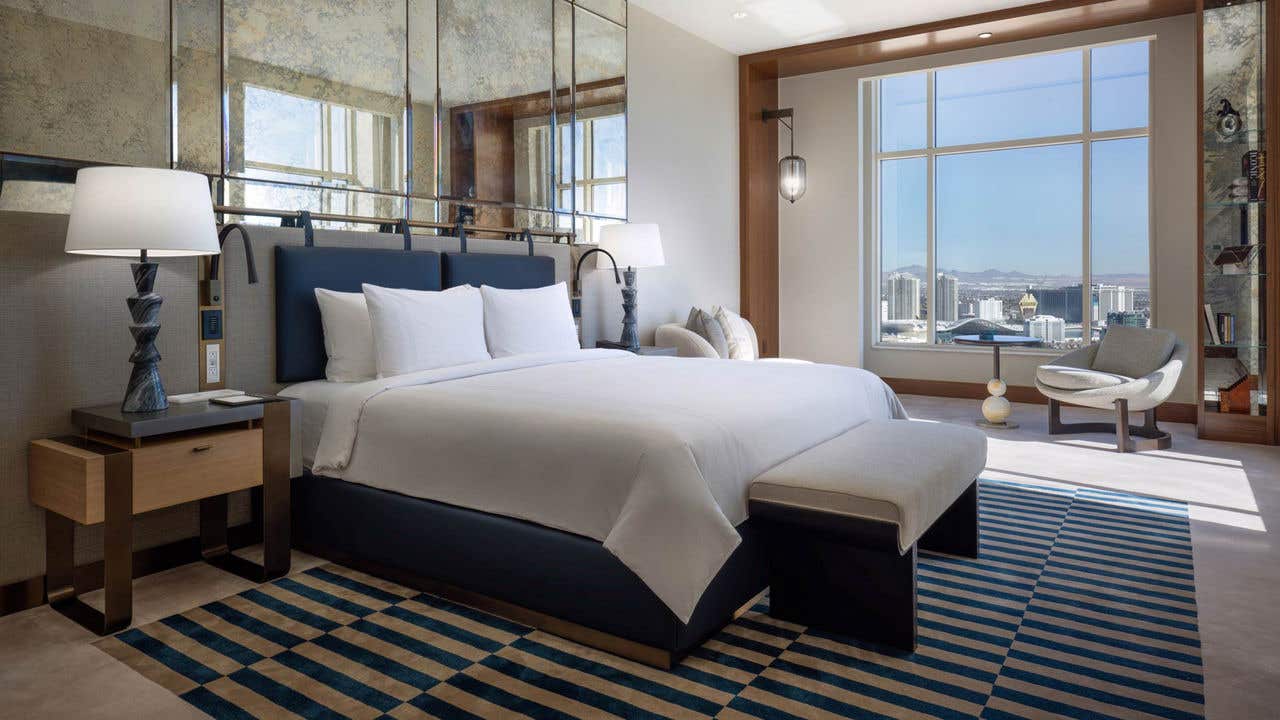 A hotel bedroom with a king bed, bench, seating area, and large window with a view of the Las Vegas Strip.