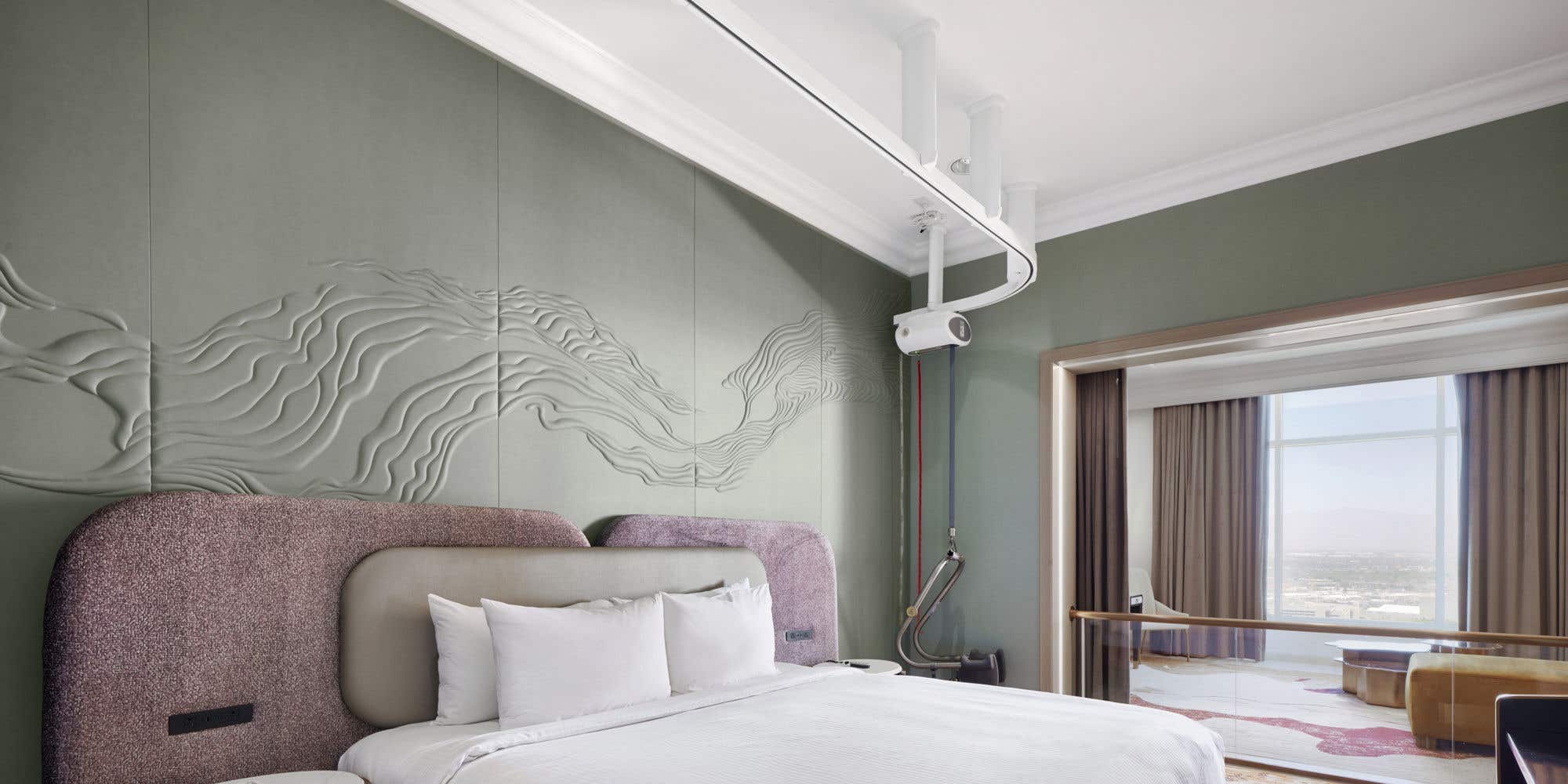 A hotel bedroom with a king bed, padded headboard, abstract wall panels, bedside tables, a mobility lift track in the ceiling going from the corner across the bed, and a glass railing separating the bedroom from the living room with a city view.