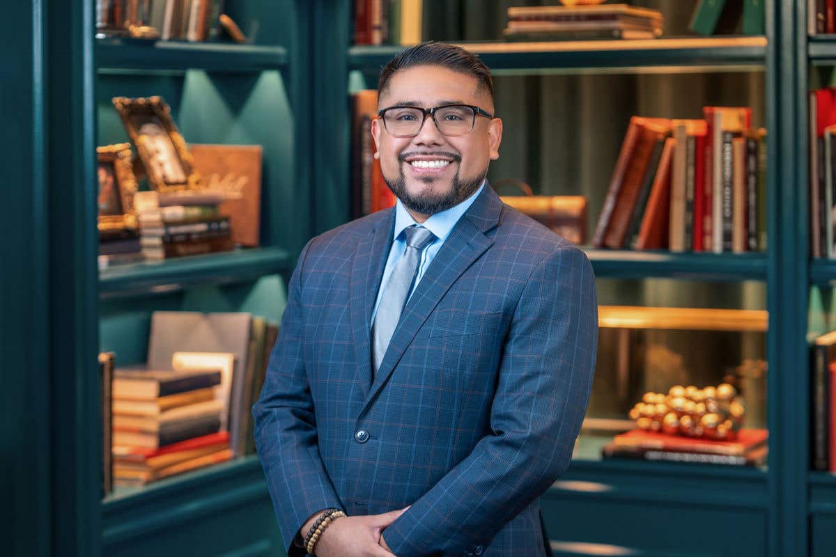 A professional headshot of a smiling man with a goatee and glasses, wearing a blue checkered suit with a grey tie. He is posed in front of a teal bookshelf background featuring colorful books and vintage decor.