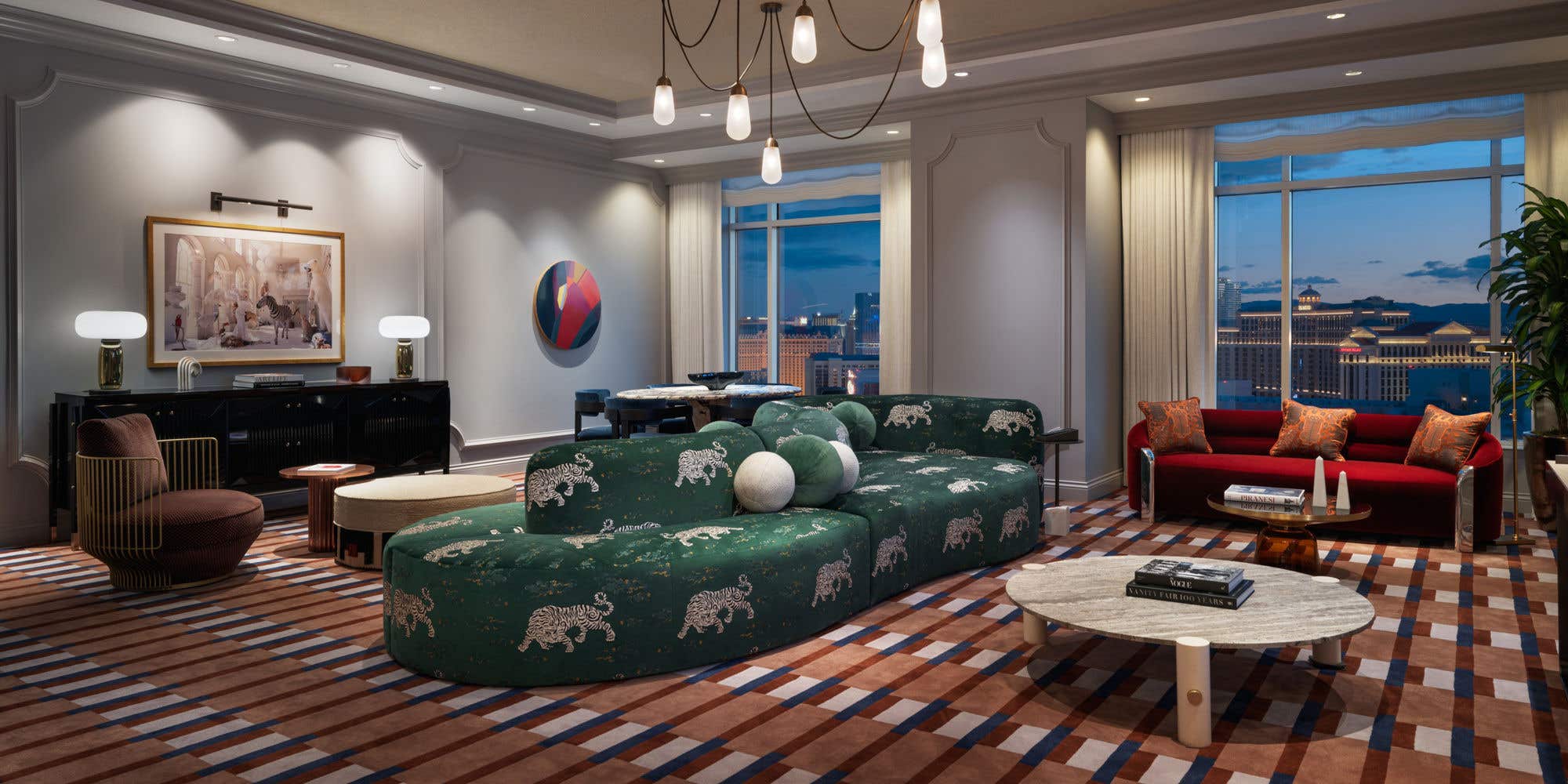 A hotel living room at night with a double-sided dark green couch, chandelier, coffee table, love seat, dining table, armchair, and two large windows with a view of the Las Vegas Strip.