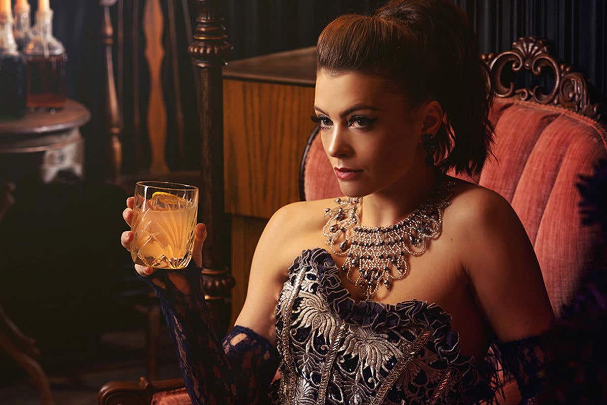 Woman sitting in a red velvet armchair dressed in glamourous attire holding a drink in her hand. 