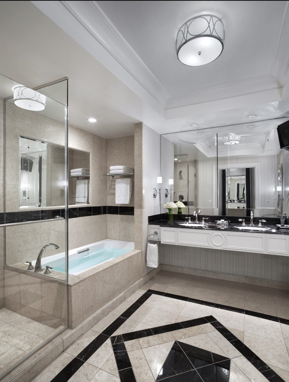 Luxurious bathroom with a bathtub, glass shower, large mirror, double sinks, and elegant black-and-white tiled flooring.