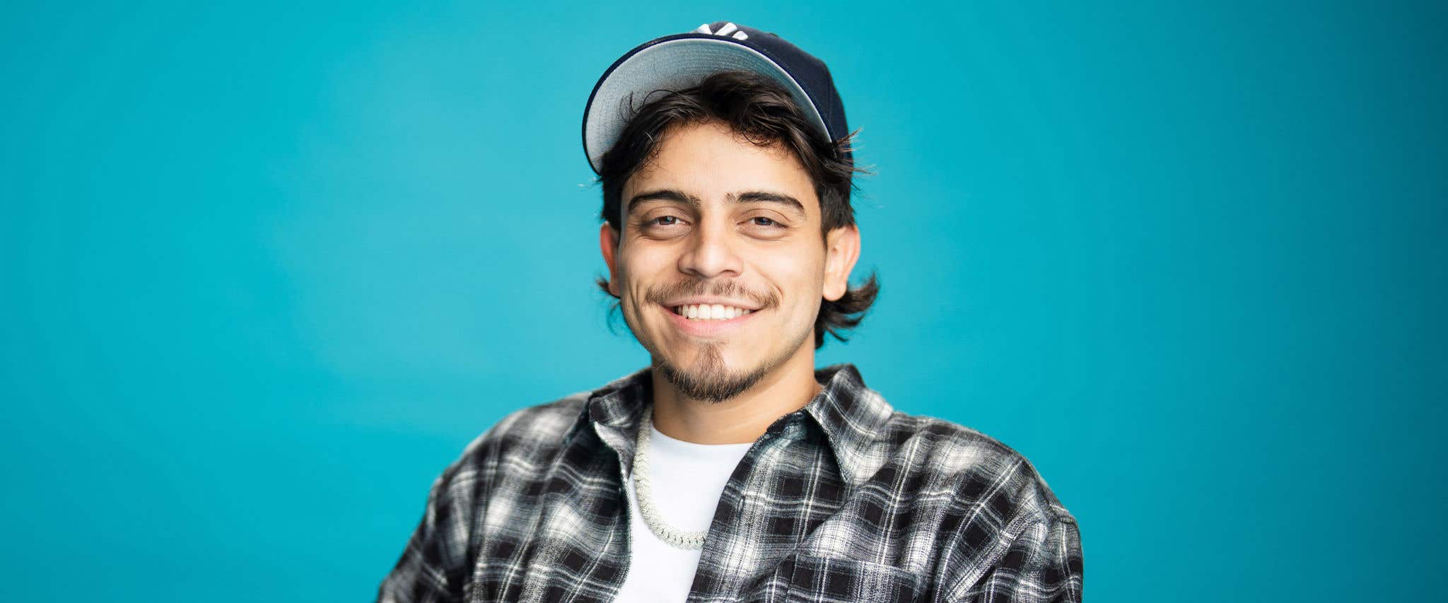 Ralph Barbosa in black and white plaid button up over white t-shirt wearing a hat and smiling at the camera. 