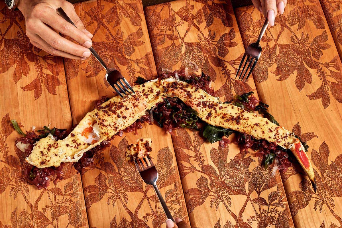 Four forks reach for a fire-roasted king crab strip on a patterned wooden table with greens and seasoning.
