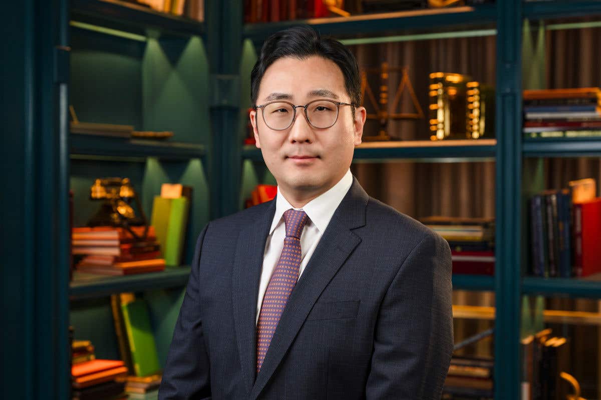 A professional headshot of a man with short black hair and glasses, wearing a dark navy suit with a white shirt and a patterned purple tie, set against a teal bookshelf background. 