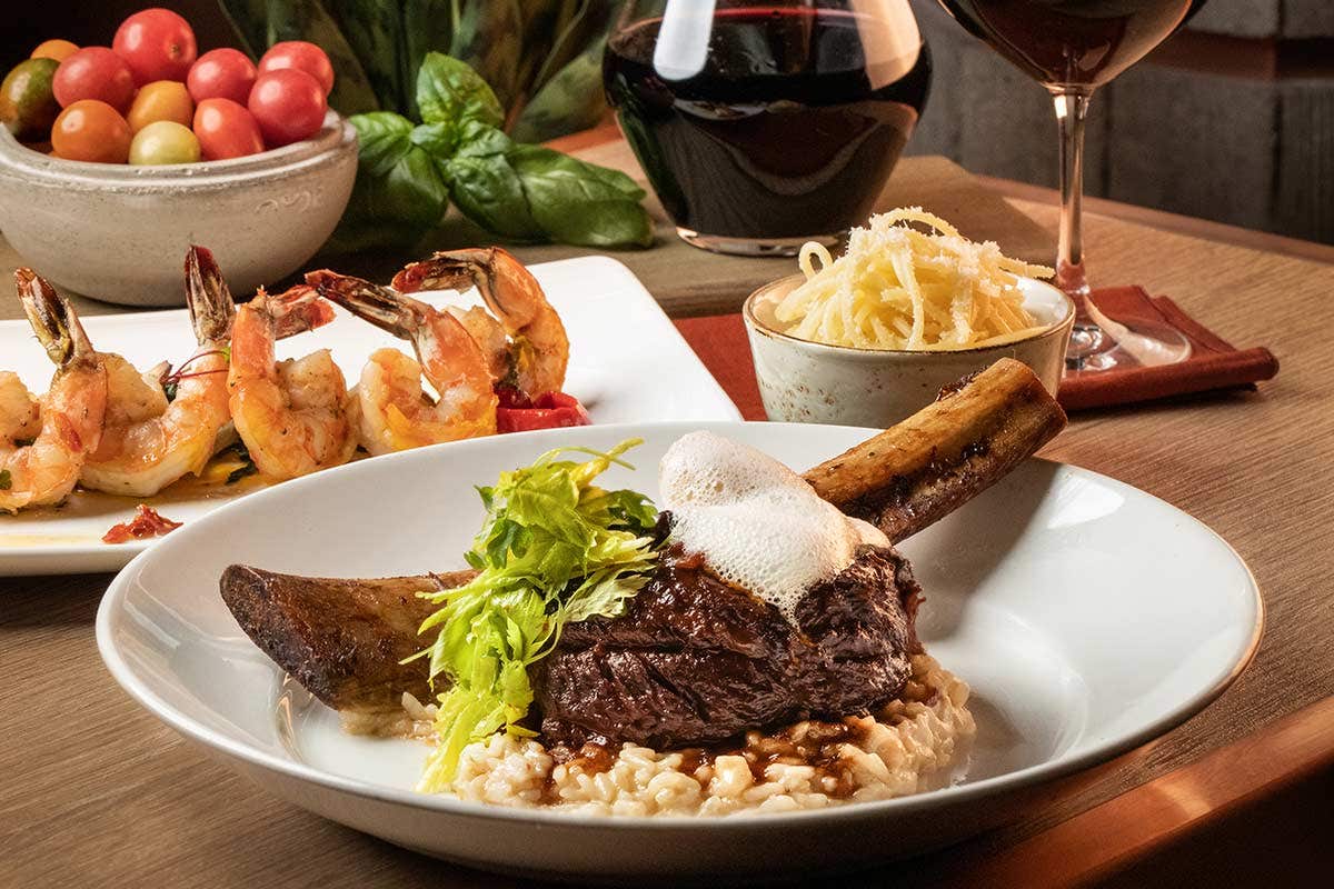 A glazed cut of beef, with the bone still attached, sits on a bed of rice and is topped with buttery foam and green lettuce. In the background is 5 roasted shrimp on a plate, and a bowl of spaghetti and parmesan, a bowl of green, orange, and red tomatoes. A pitcher and glass of red wine sit behind the food.