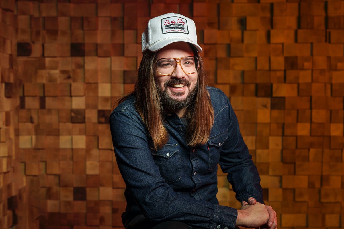 Dusty Slay with long hair and glasses, wearing a denim shirt and a white cap, smiles in front of a wooden textured background.