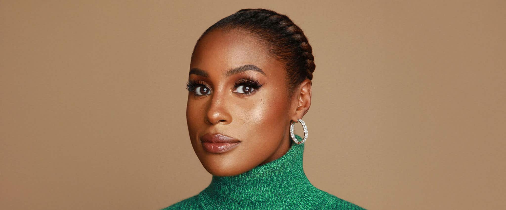 Issa Rae in a green turtle neck standing in front of beige background looking at the camera. 