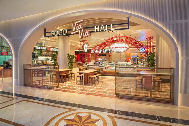 Opening to a food hall with tables, chairs and multiple restaurant fronts with a Via Via Food Hall sign in front.