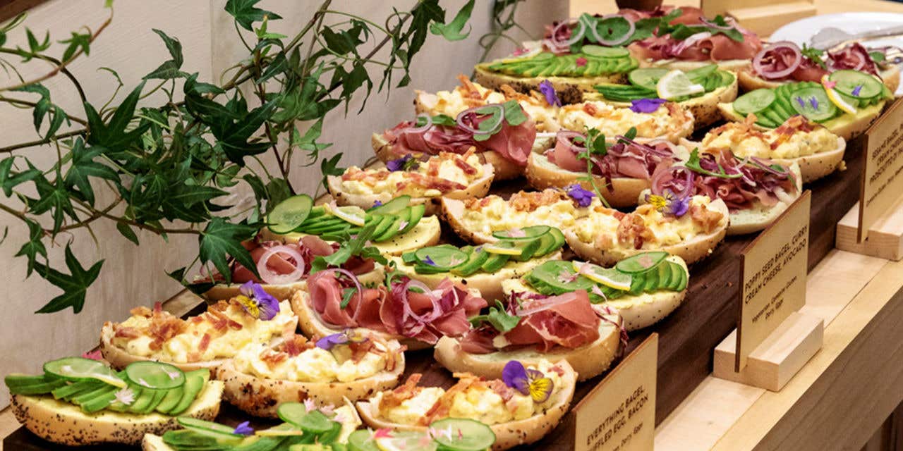 Open-faced bagel sandwiches topped with avocado, cured meat, egg salad, cucumbers, and edible flowers, arranged on wooden serving boards at a buffet.