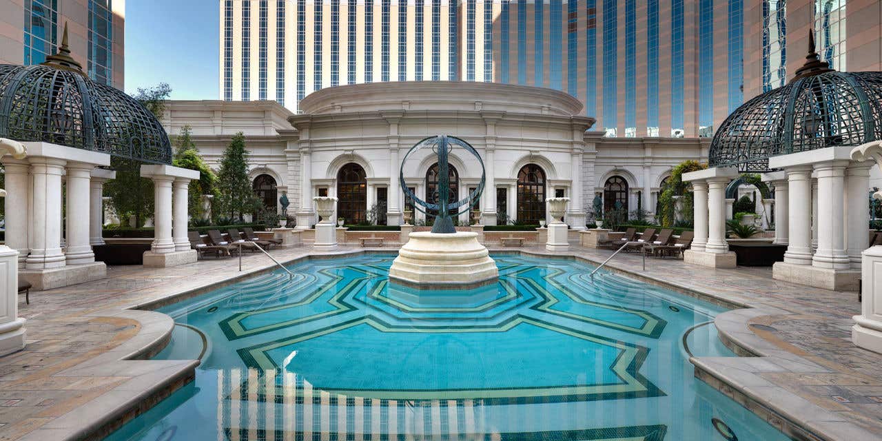 Outdoor resort pool with geometric tile patterns leading to a central circular modern sculpture, surrounded by domed pavilion structures, stone columns, and a tall hotel tower in the background.