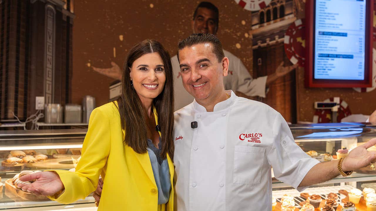 Two people pose in front of a Carlo’s Bake Shop pastry display.