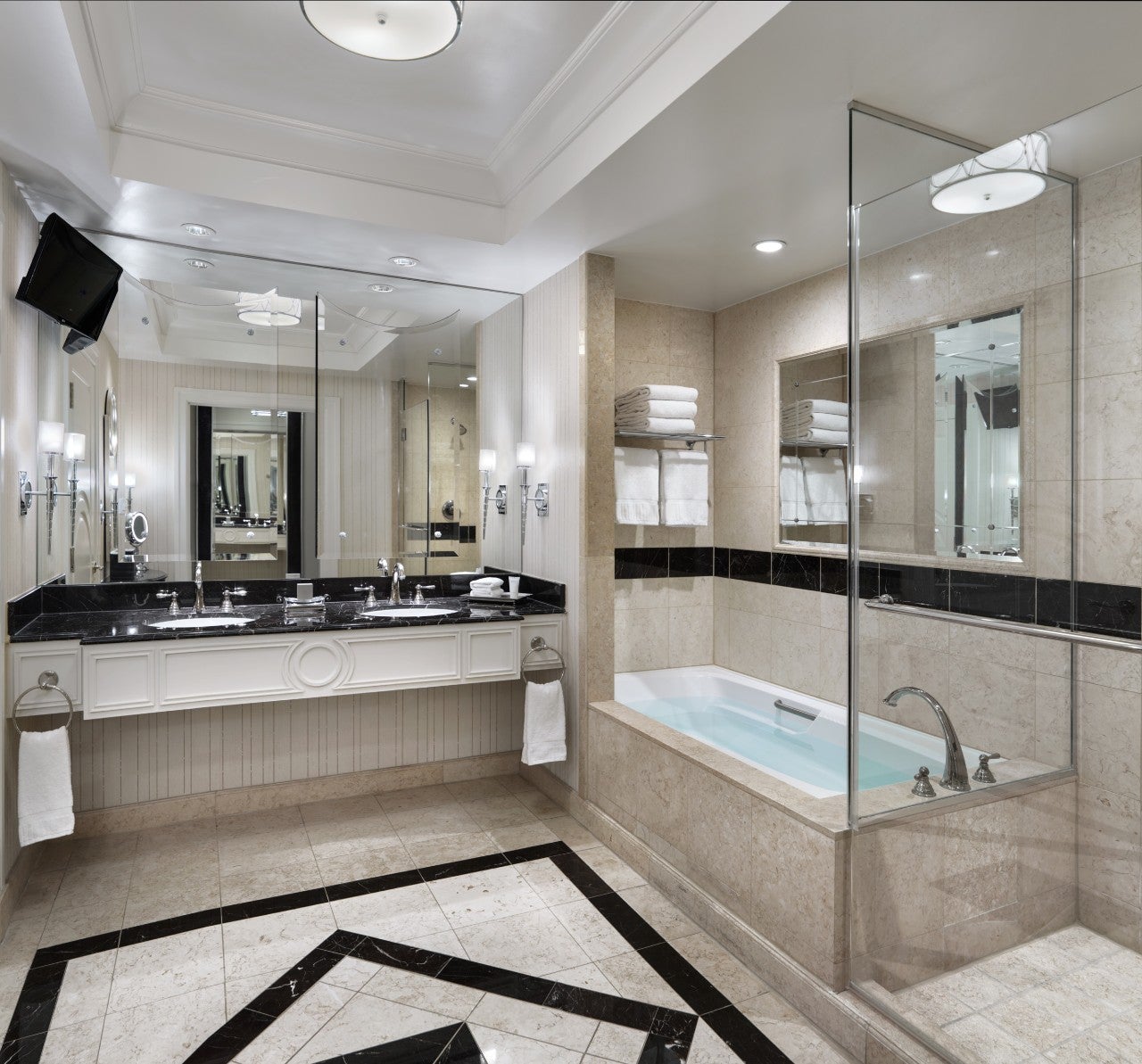 Elegant bathroom with marble tiles, double sink vanity with mirrors, bathtub, glass shower, and TV mounted on the wall.