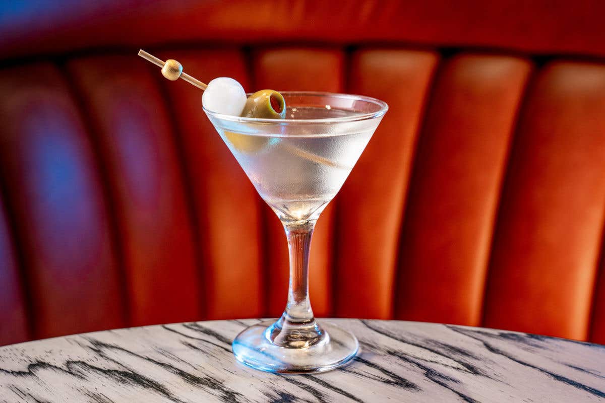 Classic martini served in a chilled cocktail glass, garnished with green olives and a pearl onion on a skewer, placed on a marble table with a tufted leather booth in the background.