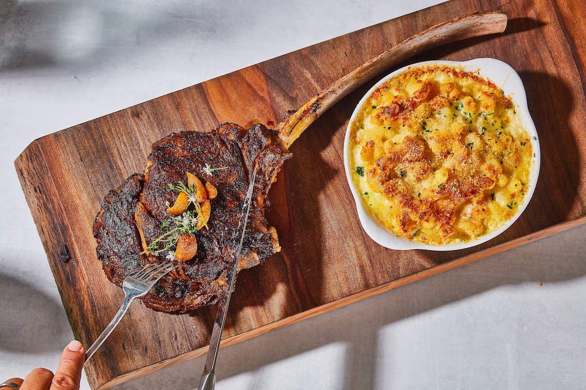 Tomahawk steak on a wooden board with a side of cheesy casserole in a white dish, and a person cutting the steak with a fork and knife.