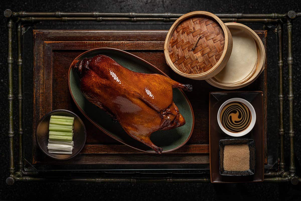 A roasted duck on a platter, bamboo steamer, dipping sauces, and sliced vegetables on a wooden tray.