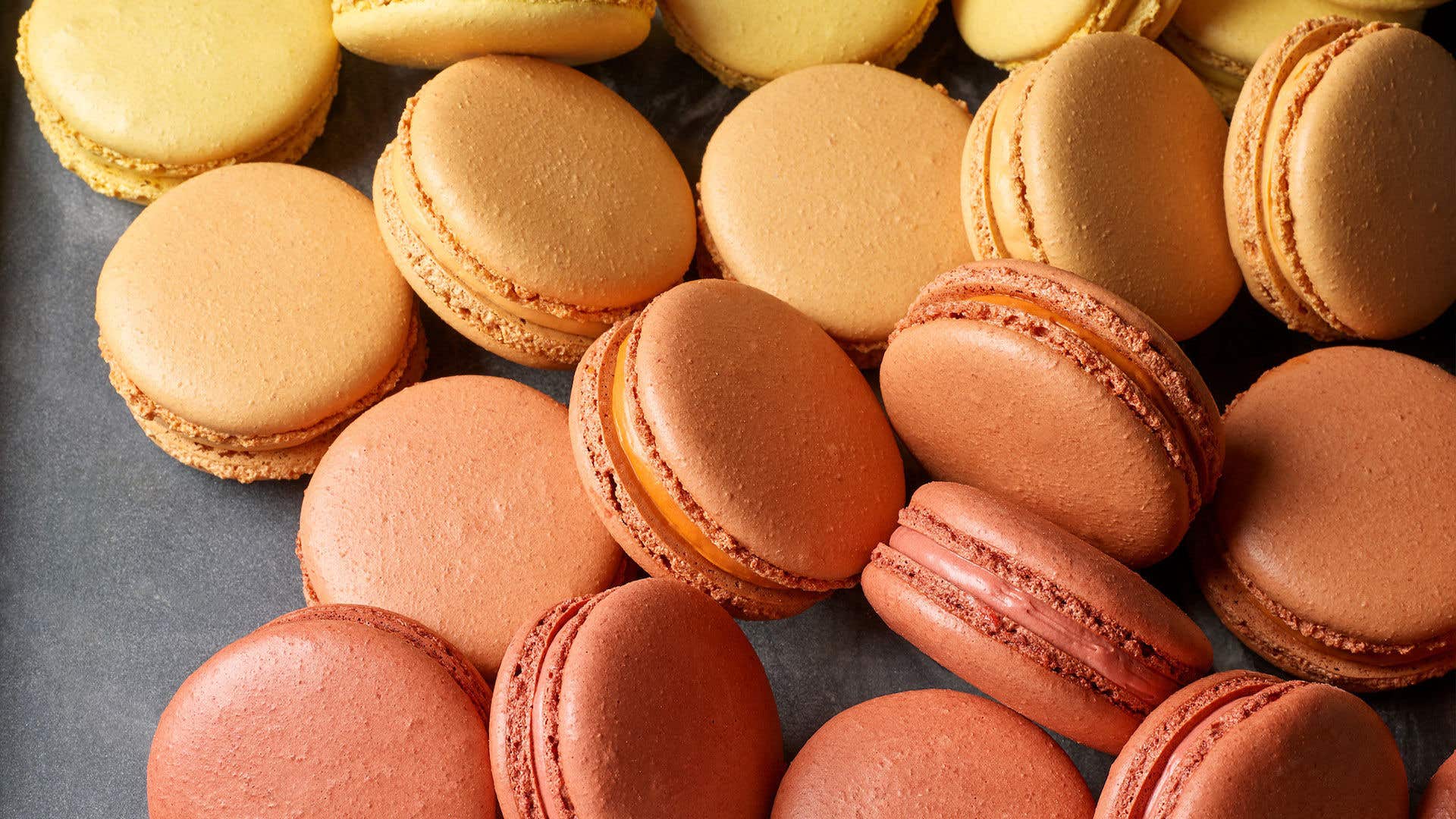 A close-up of French macarons in warm tones of yellow, orange, and rose piled on a dark tray.