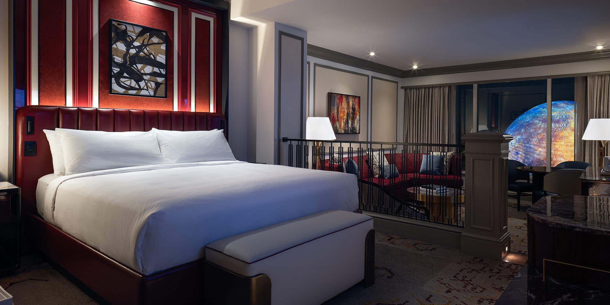 Luxury King Suite bedroom with a bench seat at the foot of the bed. In the background is a sunken living room with an armchair, couch, coffee table, dining table with chairs, lamps, and a nighttime view of Sphere at The Venetian outside the window.