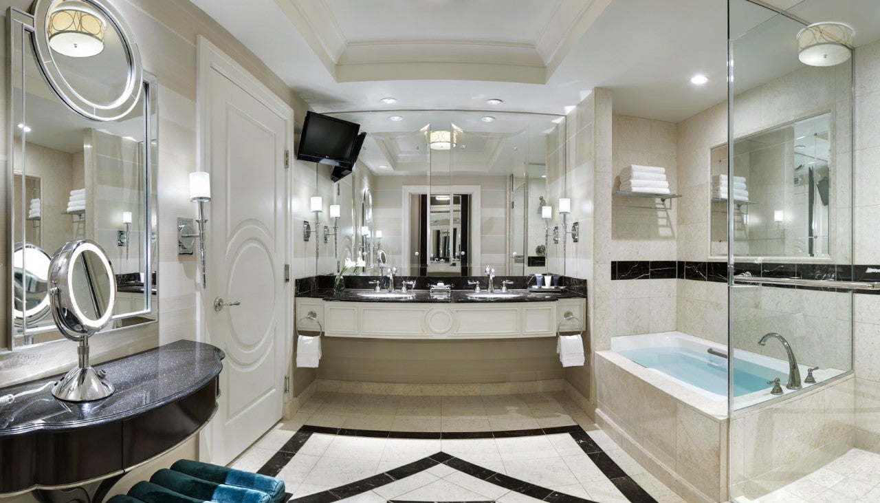 Luxurious bathroom with double sinks, a bathtub, large mirrors, towels, and a wall-mounted TV. Beige and black decor.