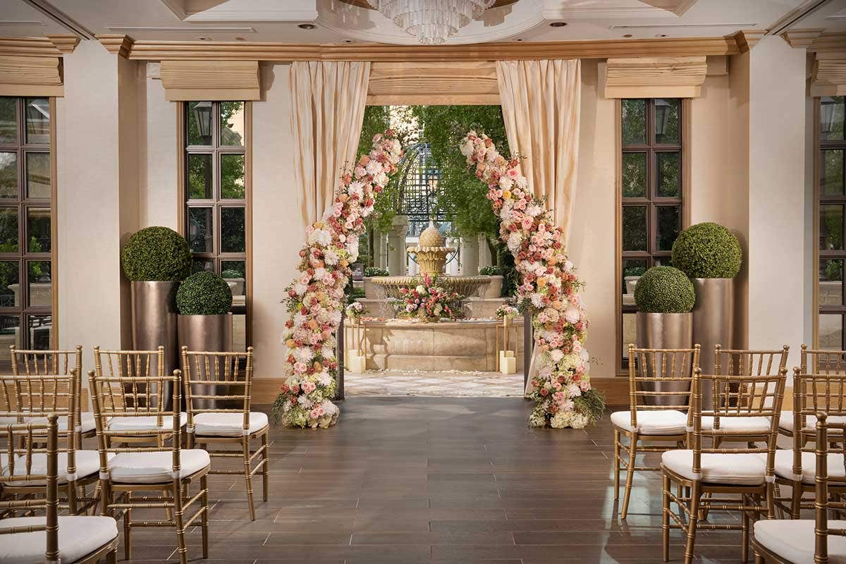 A wedding venue with chairs set up on either side of an aisle and a floral arch in front of a set of open doors looking out onto a fountain with more flowers.