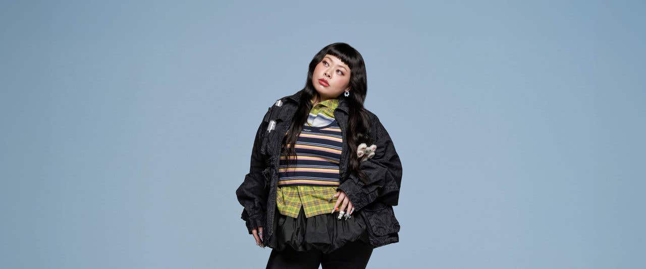 Naomi Watanabe in black jacket and bottoms with striped shirt standing in front of gray background looking off to the distance. 