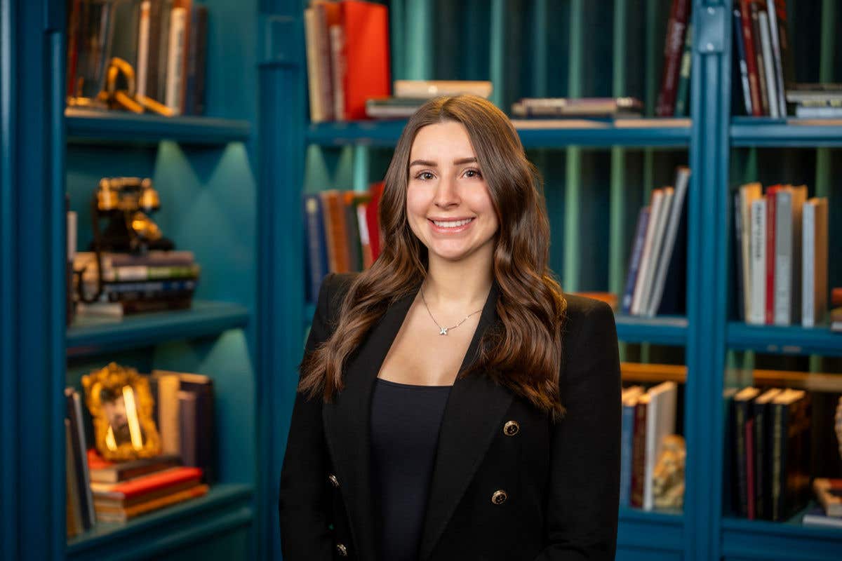 A professional headshot of a smiling woman with long brown hair, wearing a black double-breasted blazer with gold buttons and a delicate necklace, set against a teal bookshelf background.
