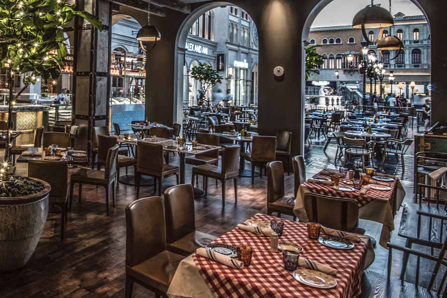 An elegant restaurant with wooden tables, checkered tablecloths, dim lighting, and a view of a brightly lit mall.