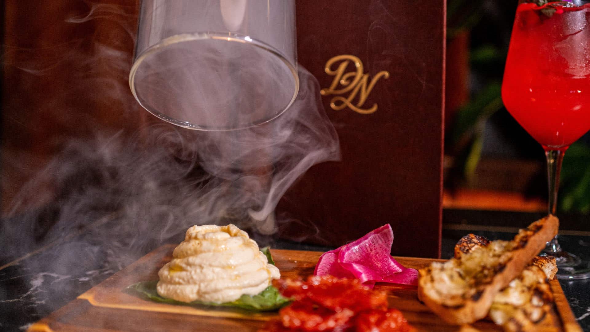 Smoked ricotta appetizer served on a wooden board with crostini and a cocktail.