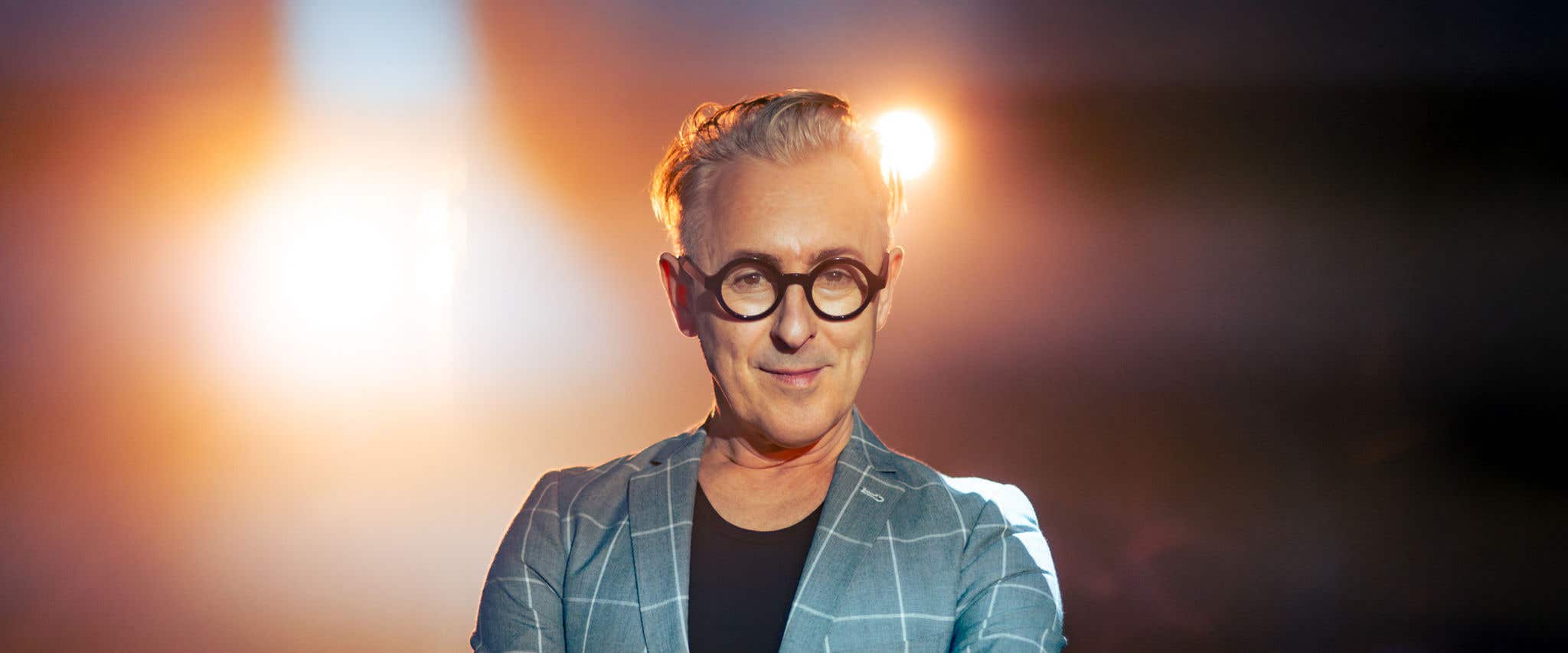 Alan Cumming wearing dark circle framed glasses looking at the camera with light shining from behind him. 