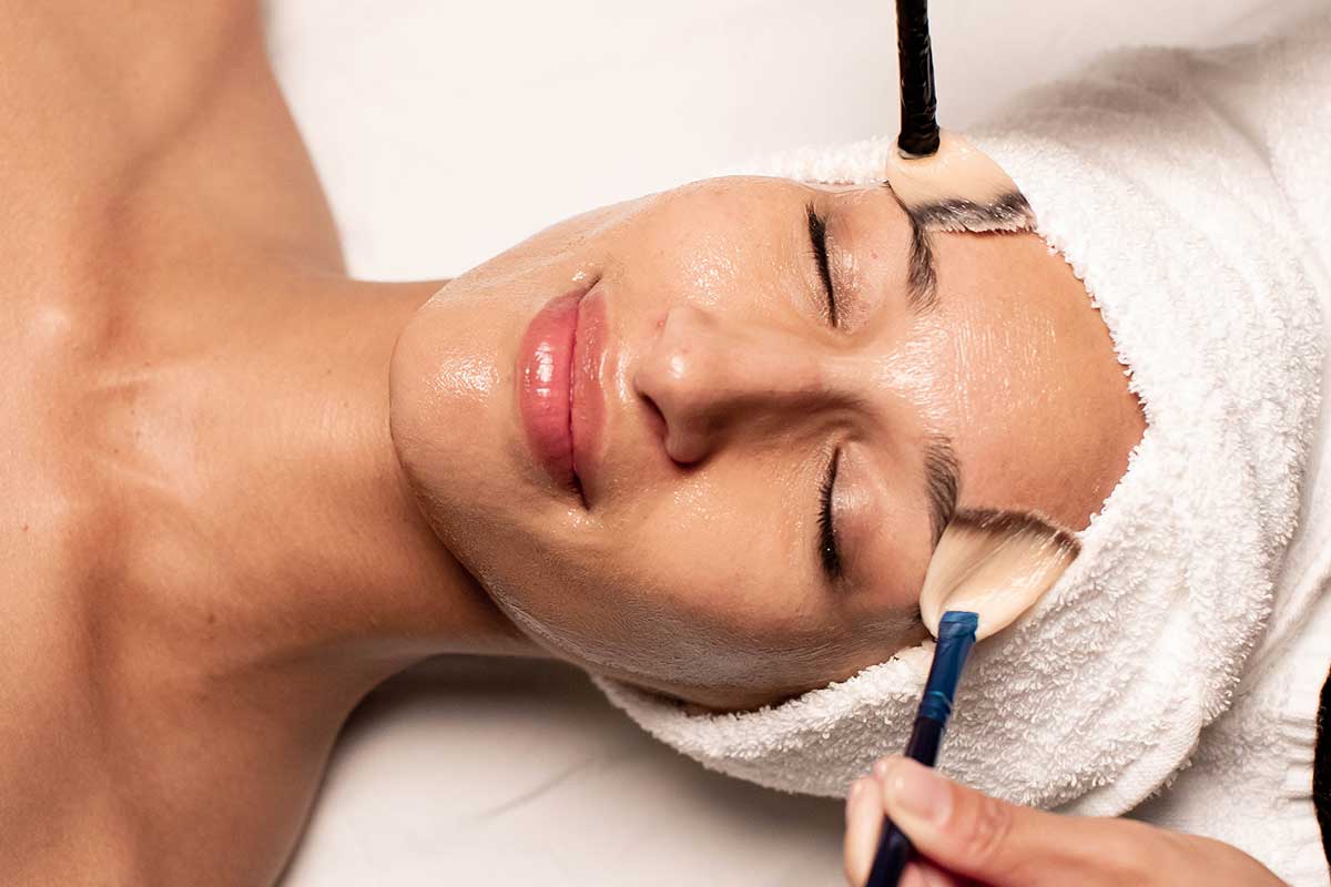 A woman lies face up with her eyes closed and hair wrapped in a towel as a facial is applied to her face with two brushes.