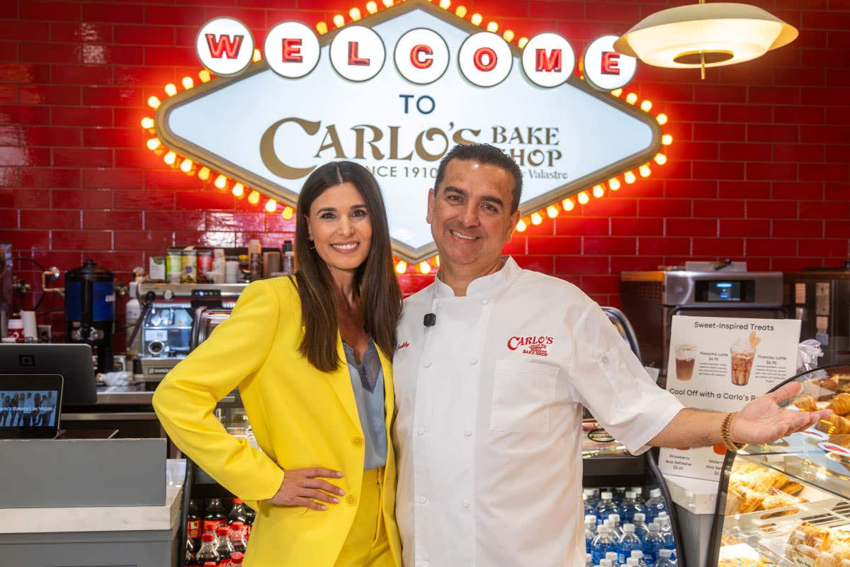 Two people pose inside Carlo’s Bake Shop beneath a bright welcome sign.