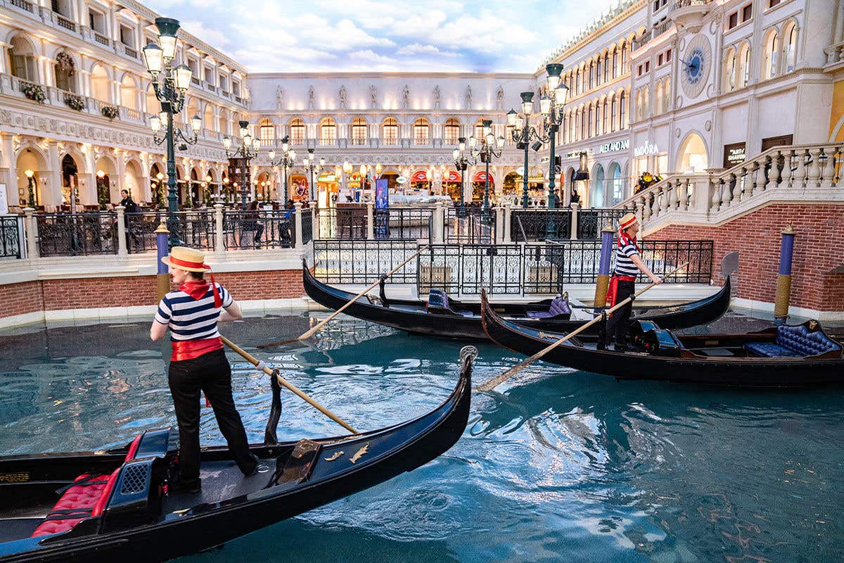 Gondoliers in traditional attire guide their gondolas through the indoor canal at The Venetian Resort Las Vegas. The waterway is lined with detailed replicas of Venetian buildings and shops, all under a ceiling painted to resemble a bright sky.