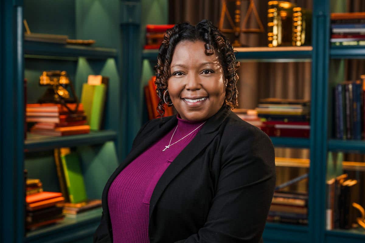 A professional headshot of a smiling woman with dark, curly hair. She is wearing a black blazer over a magenta-colored top and a delicate necklace. She is posed against a teal bookshelf background filled with books and vintage decor.