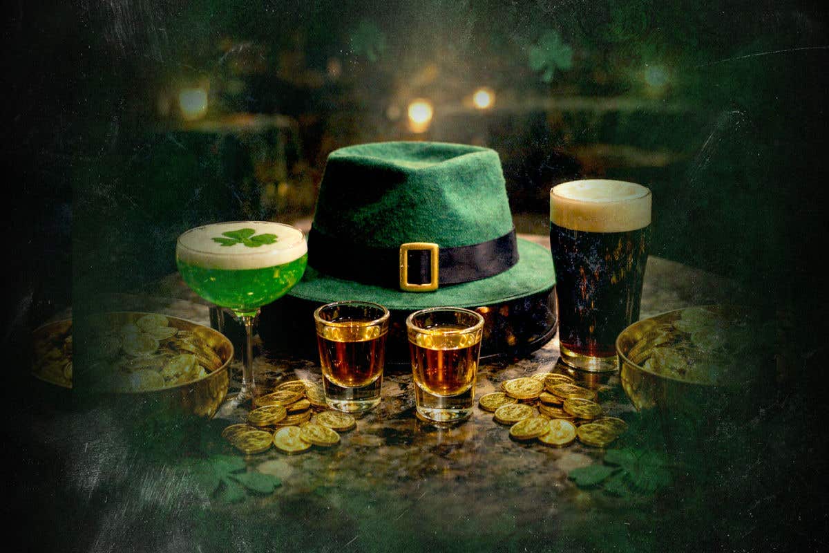 Two shots of brown liquor on a table with a green shamrock cocktail, dark beer, gold coins and a green leprechaun hat.