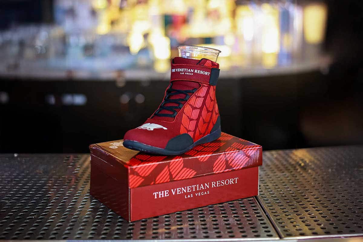 Red racing shoe with The Venetian Resort Las Vegas logo with a plastic cup sitting inside of it, resting on a shoe box on a bar