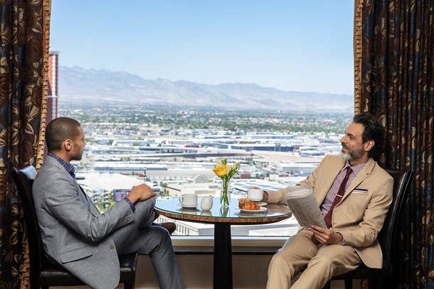 Two men in suits having a conversation over coffee and pastries at a table with a city and mountain view in the background.
