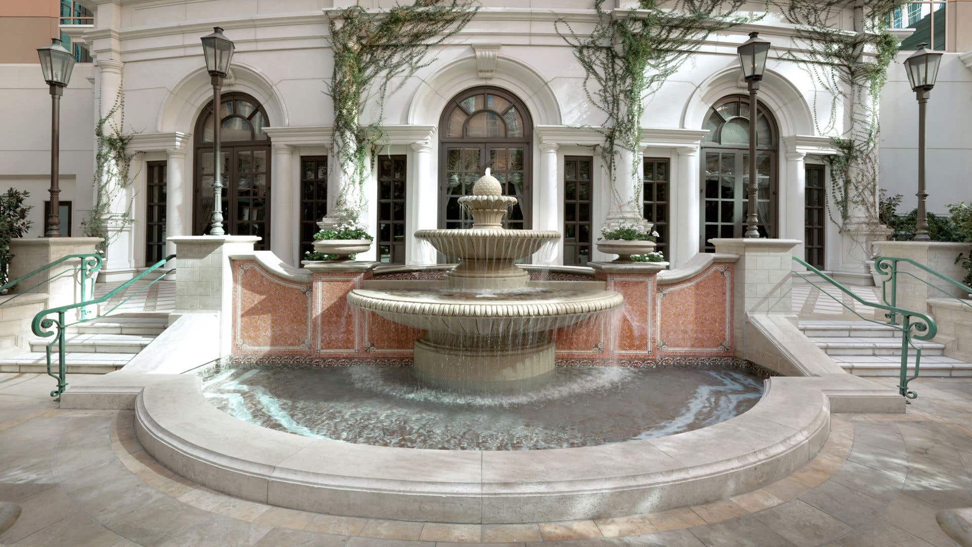 Outdoor courtyard with a large tiered stone fountain centered in front of an arched building façade, surrounded by climbing greenery, decorative lantern posts, and stairways with green handrails.
