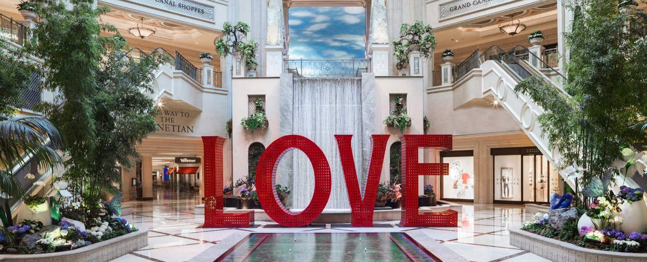 Large red "LOVE" sculpture in front of a indoor waterfall at a grand shopping mall entrance with elegant decor and greenery.
