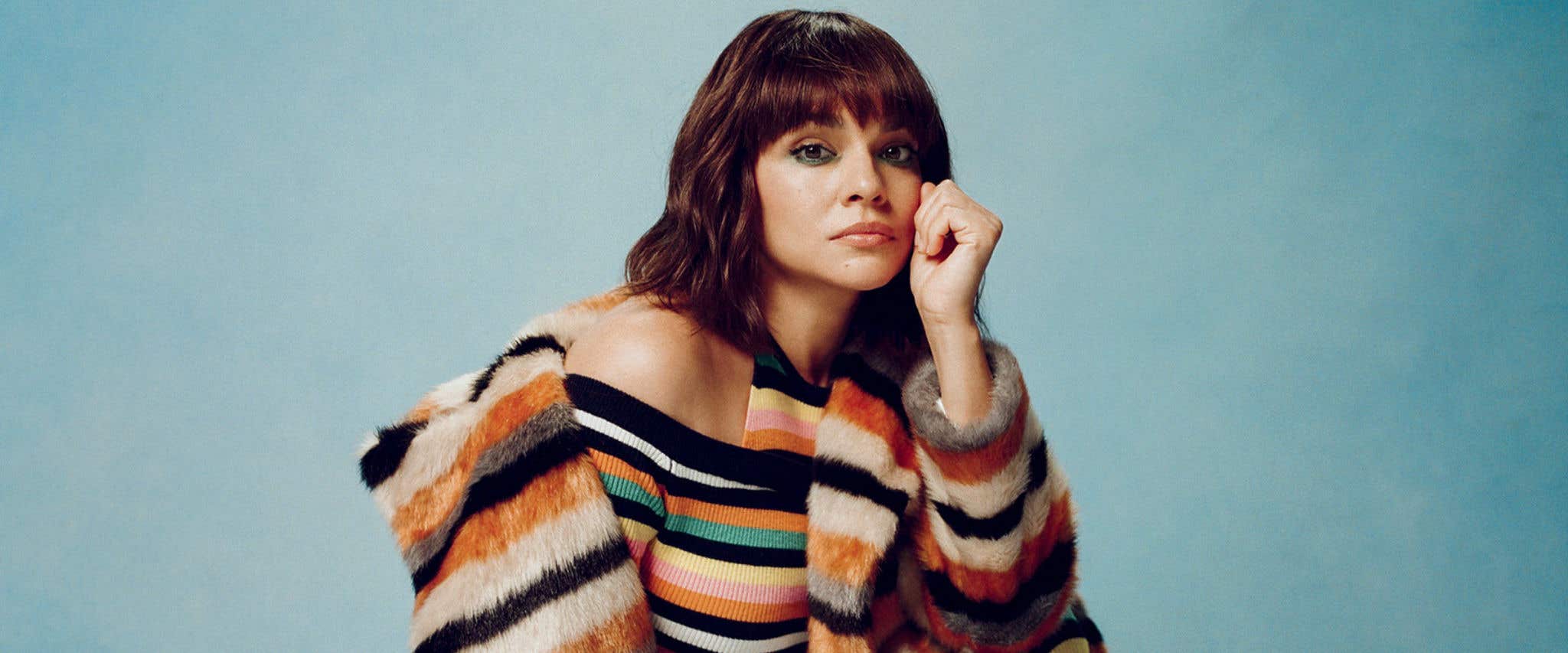 Norah Jones in a colorful striped dress and a neutral colored striped jacket posing in front of a blue background starting at the camera.