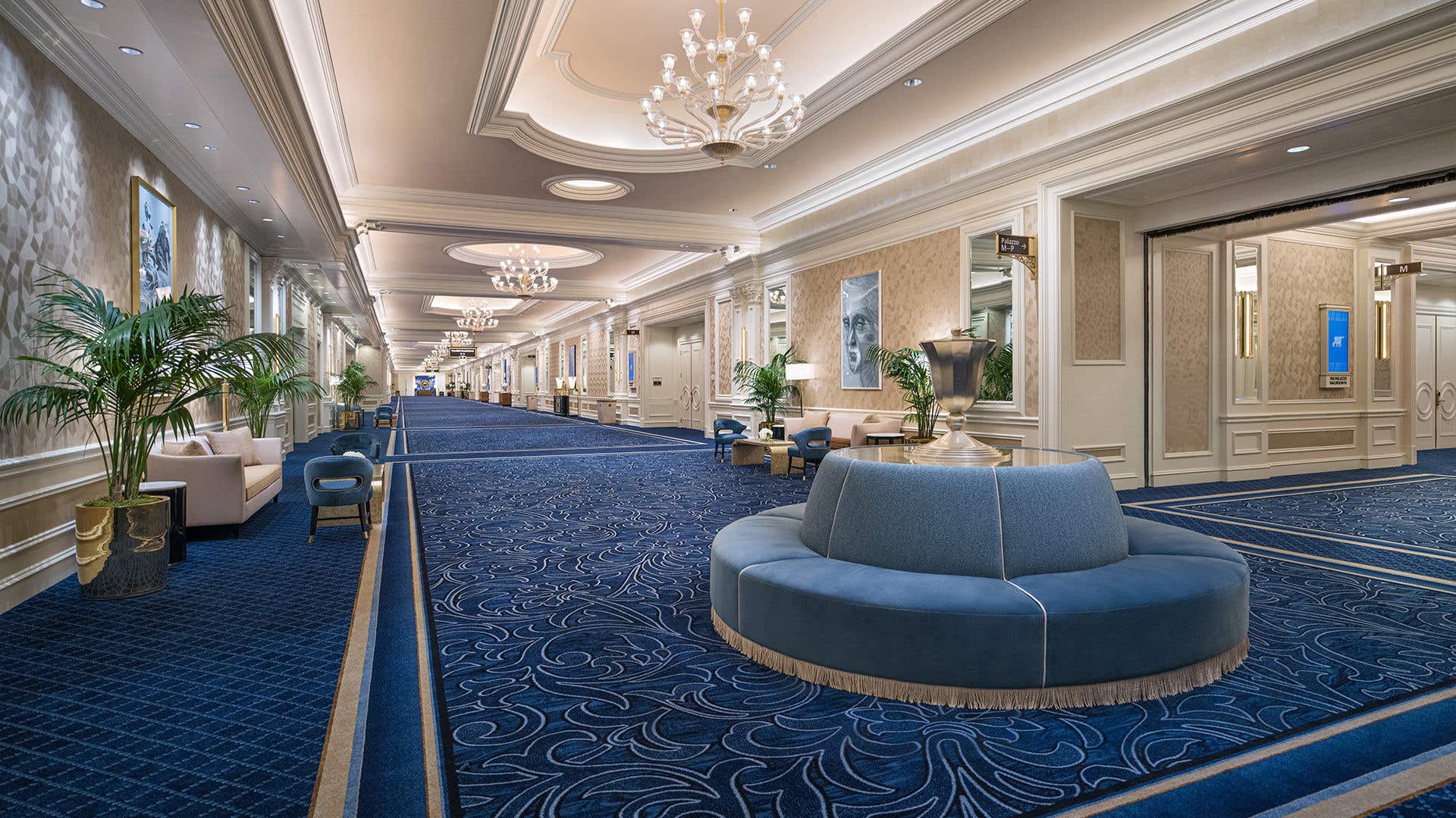 Elegant corridor with blue patterned carpet, chandeliers, ornate walls, potted plants, and a round upholstered seating area.