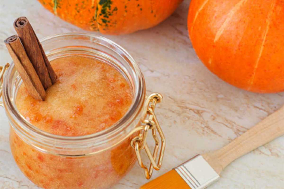A glass jar of cinnamon body scrub, a paint brush, and two small pumpkins sit on a marble table.