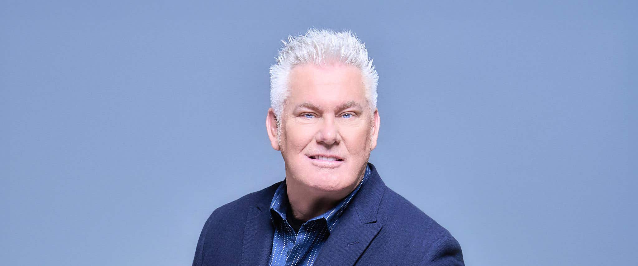 Brian Regan in a blue suit and a blue striped shirt in front of a blue background looking at the camera. 