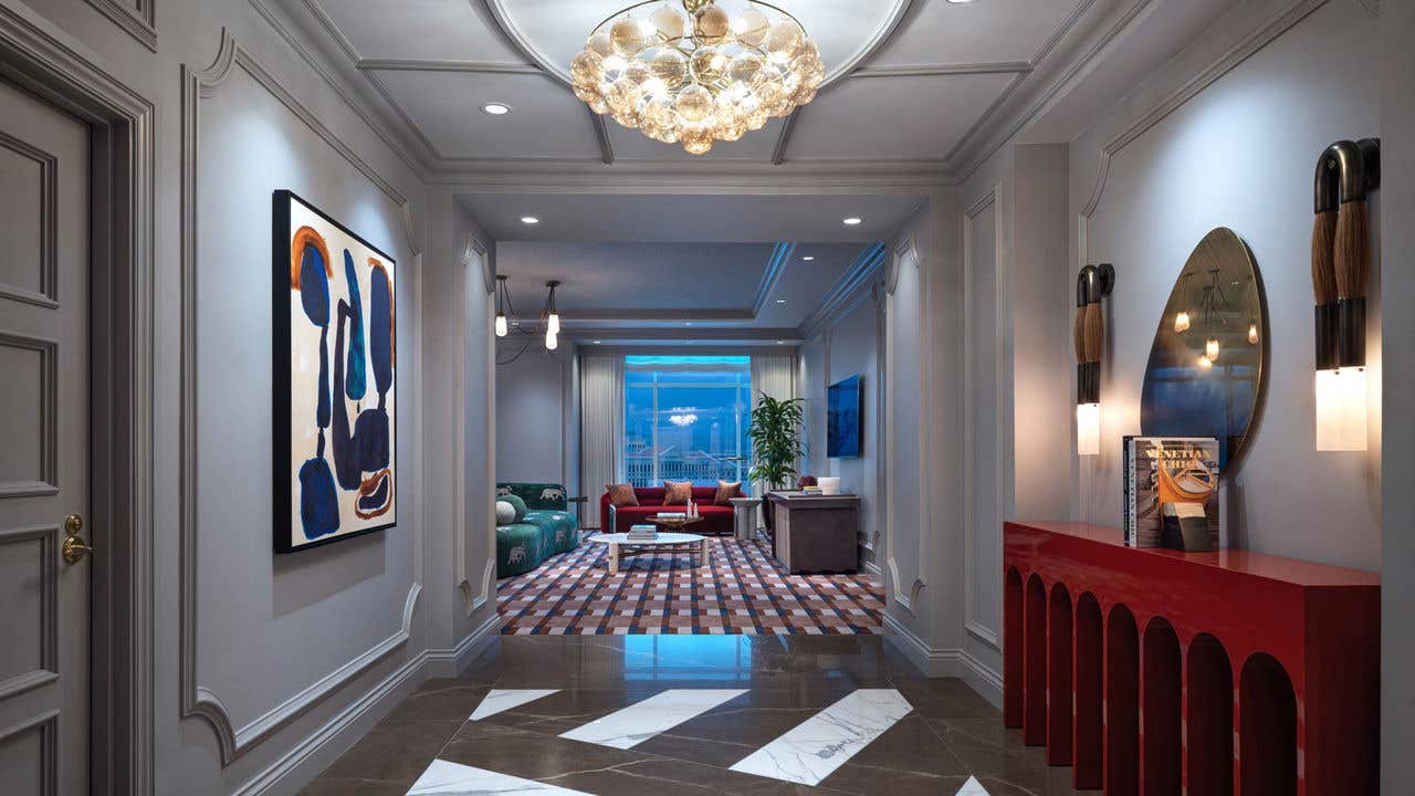 A hotel room foyer entrance with a small chandelier, wall-mounted painting on the left, a side table, mirror, and wall sconces on the right, and an open living room straight ahead.
