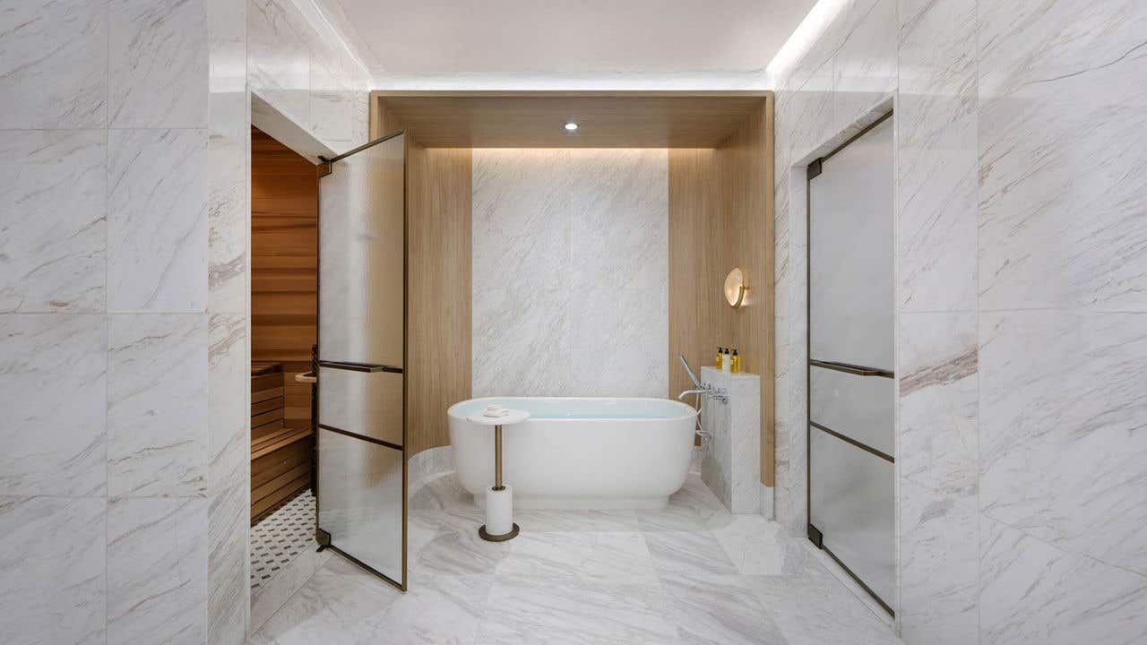 A white marble bathroom with a freestanding bathtub in the middle, frosted glass door leading to a wood sauna on the left, and another frosted glass door on the right.